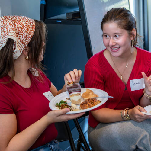 Two students share food and conversation during a College of Computing mixer at GVSU. Both are smiling and engaged, highlighting the sense of community and connection fostered at the event.